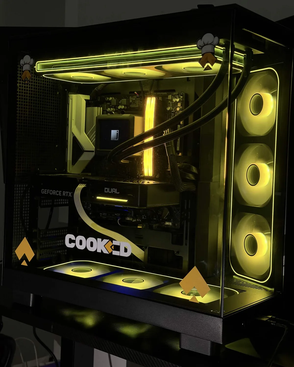 The giveaway PC on a desk: a black mid-tower with a glass side panel, lit yellow, showing a liquid-cooling radiator across the top, a GeForce RTX graphics card and a COOKED decal across the glass.